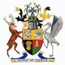 A shield in quadrants has a sheaf of wheat, the heads of a bull and ram, and a column of gold rising from a heap of quartz. Above is a crest showing the state badge of the royal crown and a Maltese cross framed by two stalks of sugar cane. On either side of the shield is a red deer and a brolga.