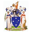 A blue shield shows the five stars of the Southern cross. It is supported by two female figures (peace and prosperity). Pink heath grows from the grassy mound below. Above the shield is a wreath of silver and blue and the torso of a Kangaroo holding a crown.